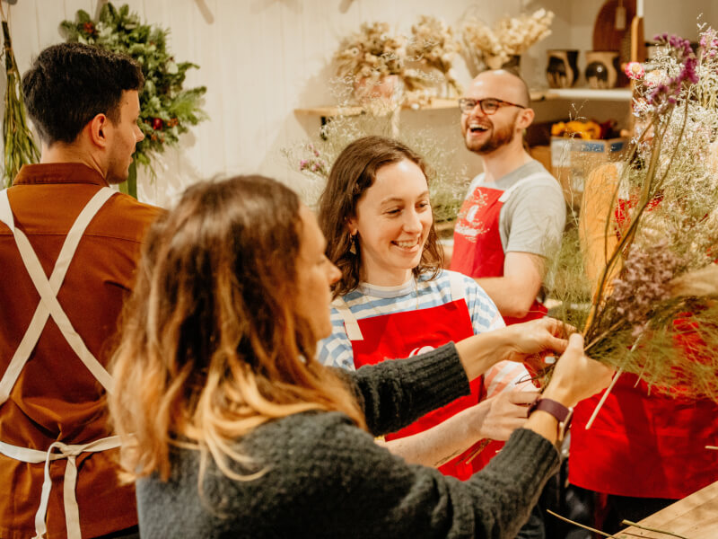 Two women are arranging a bouquet of dried flowers, with two men in the background